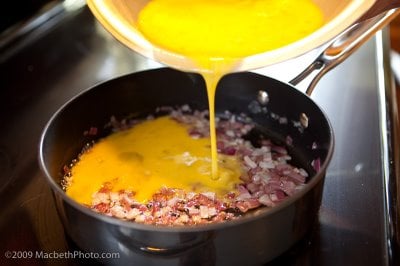 Pouring scrambled eggs into frying pan with sautéed onions.