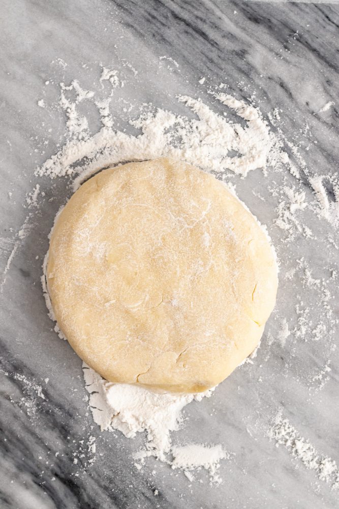 Overhead view of dough on floured marble surface formed into a flat disc shape.