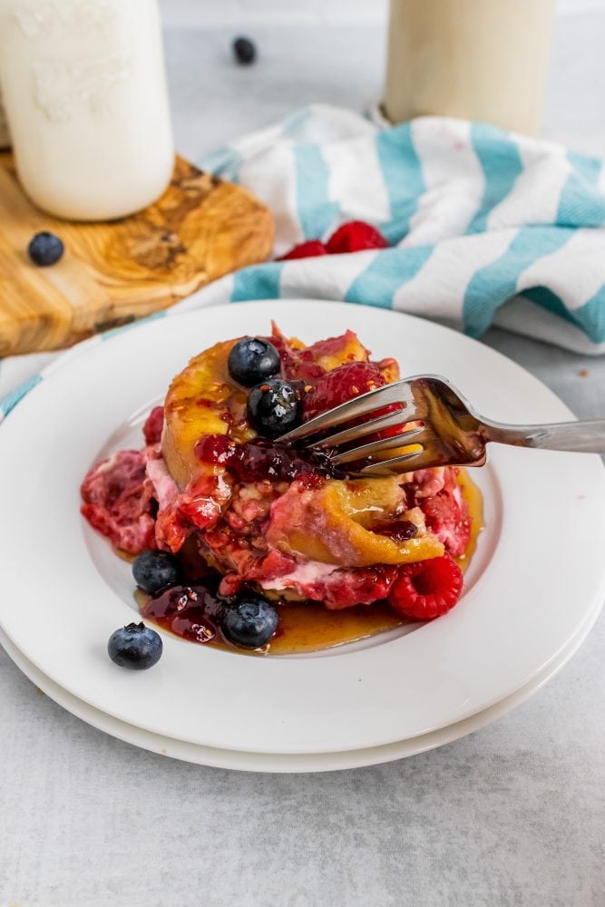 Side view of a fork cutting into a first bite of cream cheese stuffed french toast topped with fresh blueberries and raspberries.