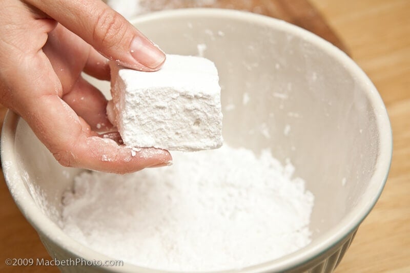 hand holding large homemade marshmallow over bowl filled with powdered sugar