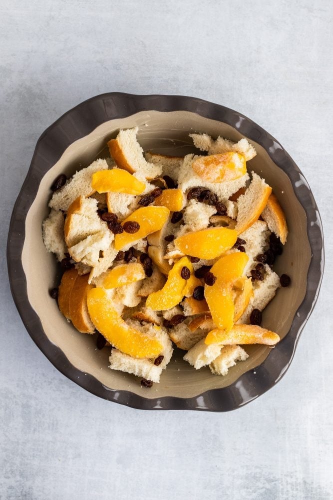 Overhead shot showing layering of bread, peaches, and raisins in a ceramic pie pan.