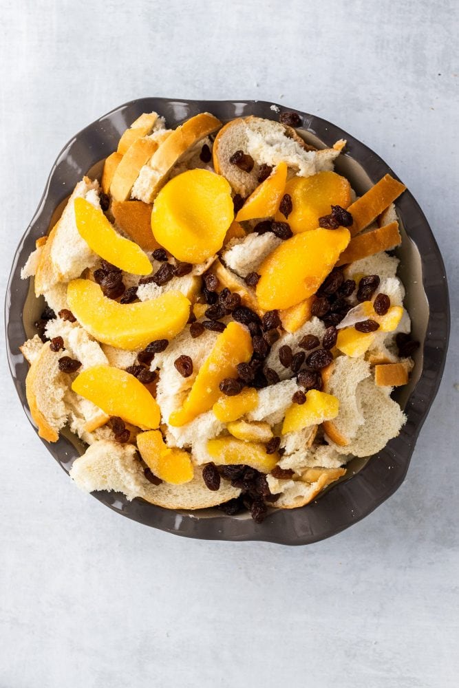 One final overhead shot showing layering of peaches, bread, and raisins on top of the cream cheese cubes. The whole bread pudding is now ready to bake.