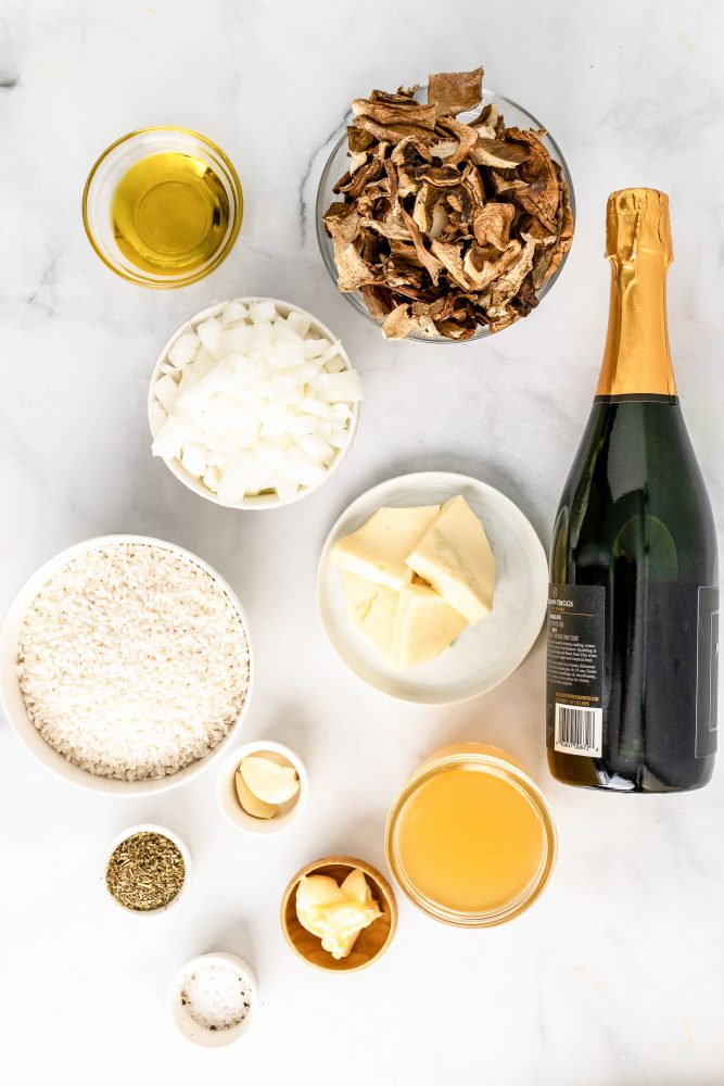 Overhead look at ingredients needed for Champagne risotto including olive oil, Champagne, dried mushrooms, diced onions, butter, short grain rice, garlic, salt, Italian seasoning, and broth.