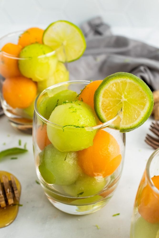 Two small glass cups with cantaloupe and honeydew melon balls topped with lime zest and garnished with a slice of lime.