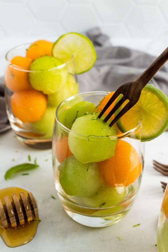 Two small glass cups with cantaloupe and honeydew melon balls topped with lime zest and garnished with a slice of lime. A small cocktail fork is digging into one of the honeydew melon balls.
