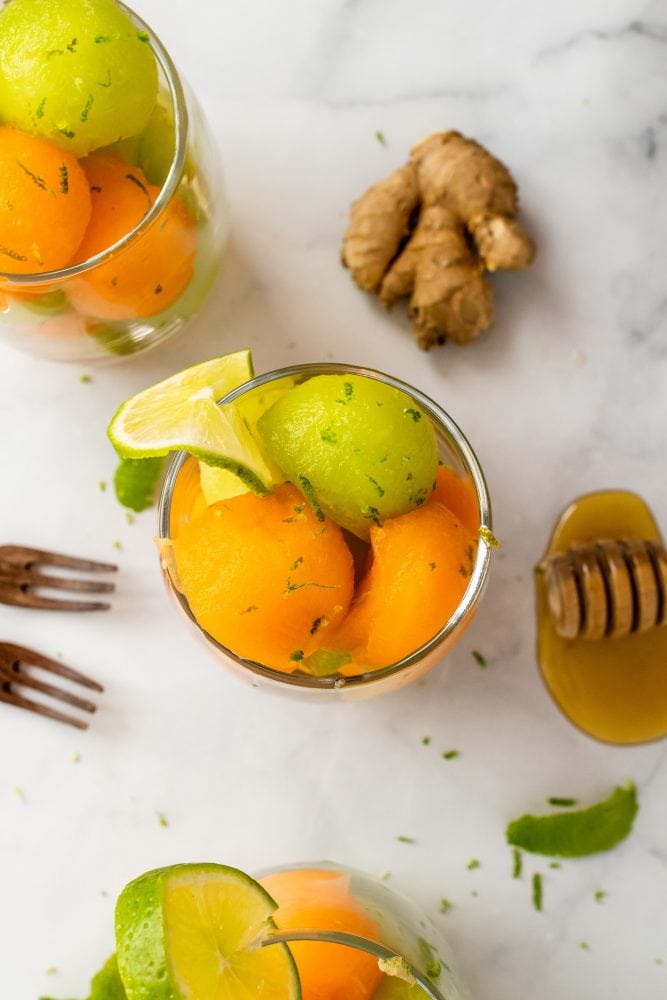 Overhead shot of three glass cups with cantaloupe and honeydew melon balls topped with lime zest and garnished with a slice of lime.