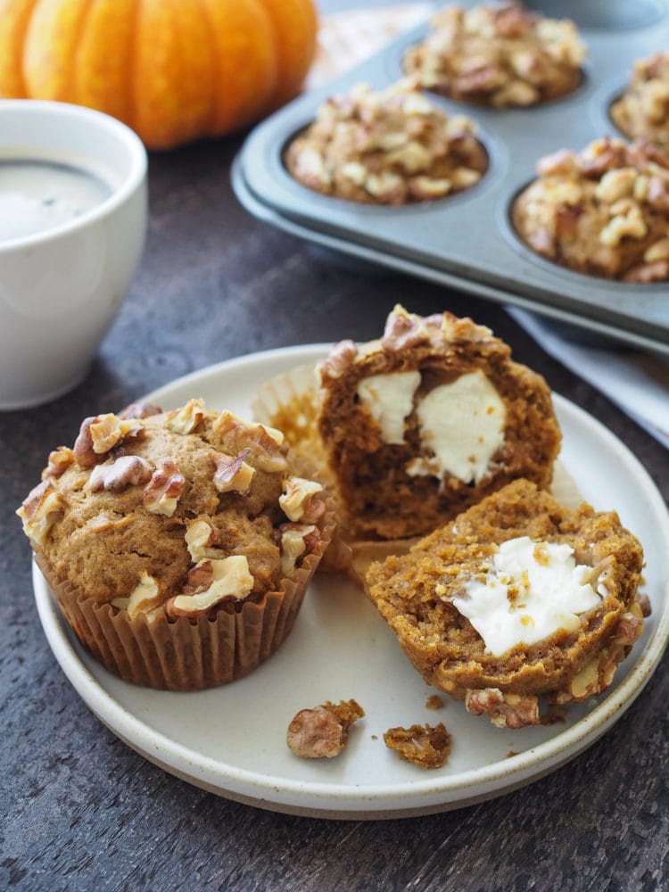 Pumpkin Cream Cheese Muffins on a plate, one is cut in half to see the cream cheese filling. And to the side there's a mini decorative pumpkin, a fork, a cup of coffee, and the edge of a muffin tin.