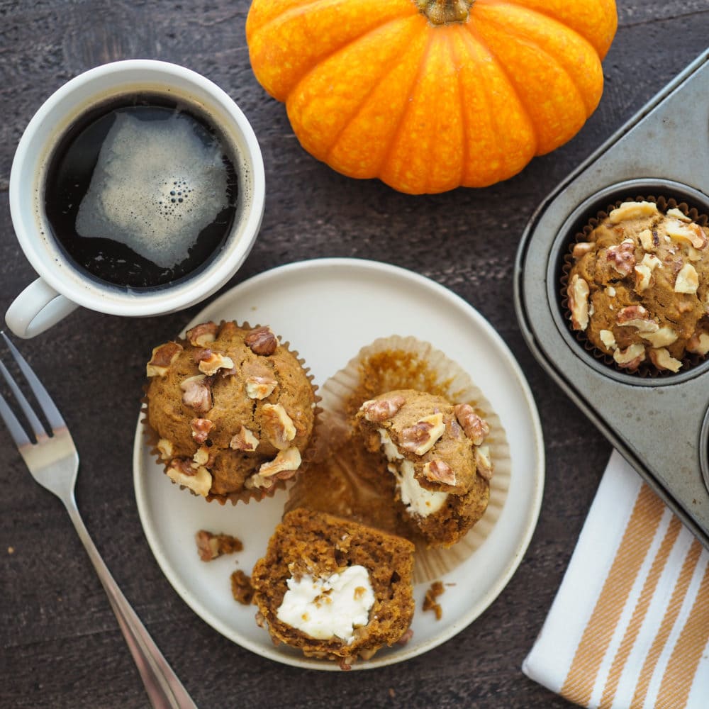 Pumpkin Cream Cheese Muffins on a plate, one is cut in half to see the cream cheese filling. And to the side there's a mini decorative pumpkin, a fork, a cup of coffee, and the edge of a muffin tin.