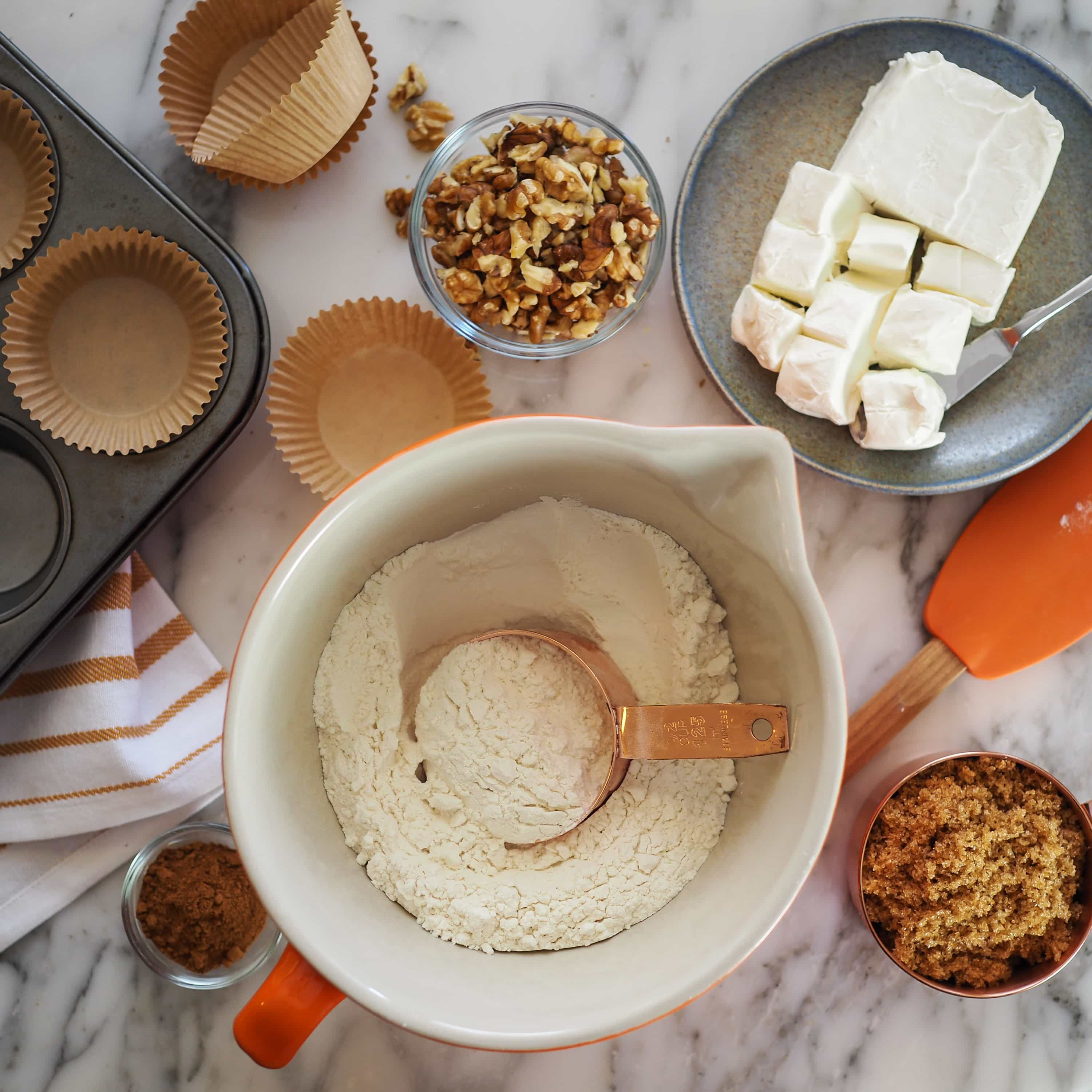 Overhead photo of kitchen table making pumpkin cream cheese muffins. There's a bowl of flour, muffin tin with tan paper muffin cups, and a block of cream cheese cubed.