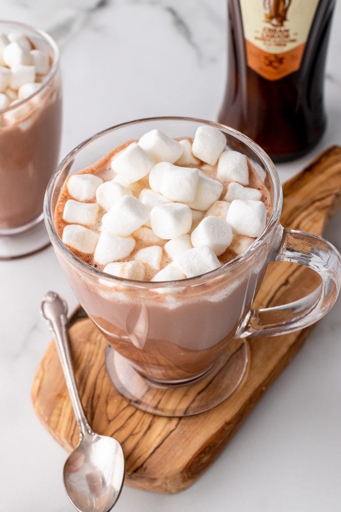 Clear glass mug filled with creamy amarula hot chocolate, topped with marshmallows, and resting on a wood board with a spoon.