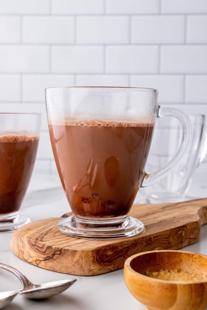 Hot chocolate in a clear mug on a wood board with a small bowl of sugar on the side.