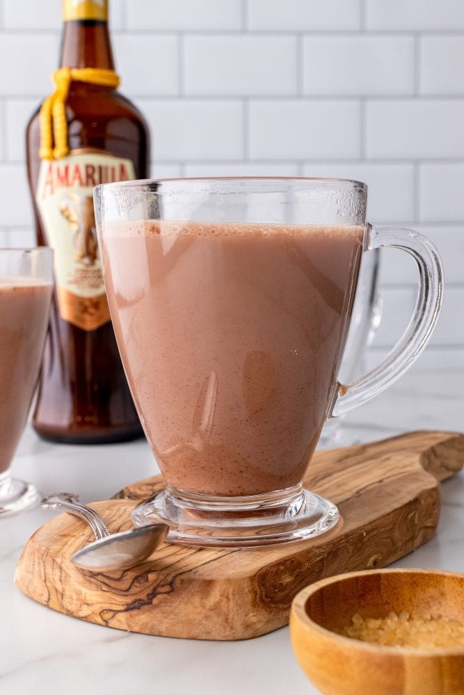 Creamy hot chocolate with Amarula in a clear mug on a wood board with a spoon and small bowl of sugar. Bottle of Amarula in background.