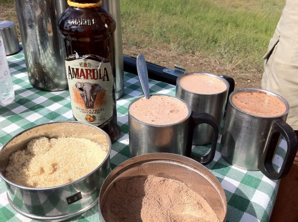 Picnic table with green and white checkered cloth holding three mugs of amarula hot chocolate, a bottle of amarula, a tin of sugar, and a tin of hot chocolate mix. 