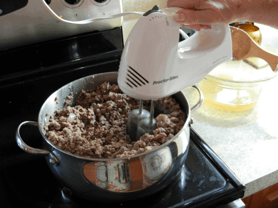 A handheld blender mixing the meat and spices in a large stainless steel pot.