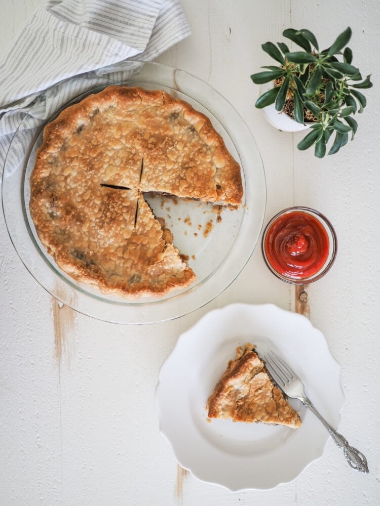 Overhead photo of a whole tourtiere, or meat pie, with a slice on a plate, a side of ketchup, and a decorative small plant.