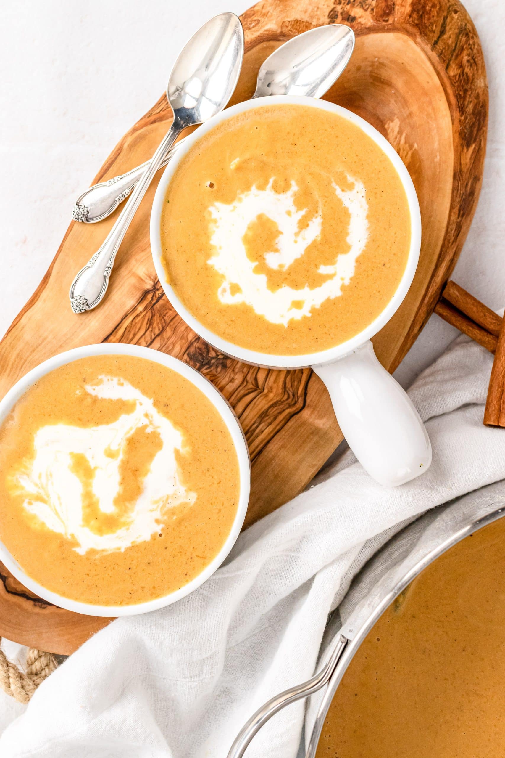 Overhead look at two bowls of butternut squash soup with swirl of cream on top. Two serving spoons and two cinnamon sticks off to the side for garnish.