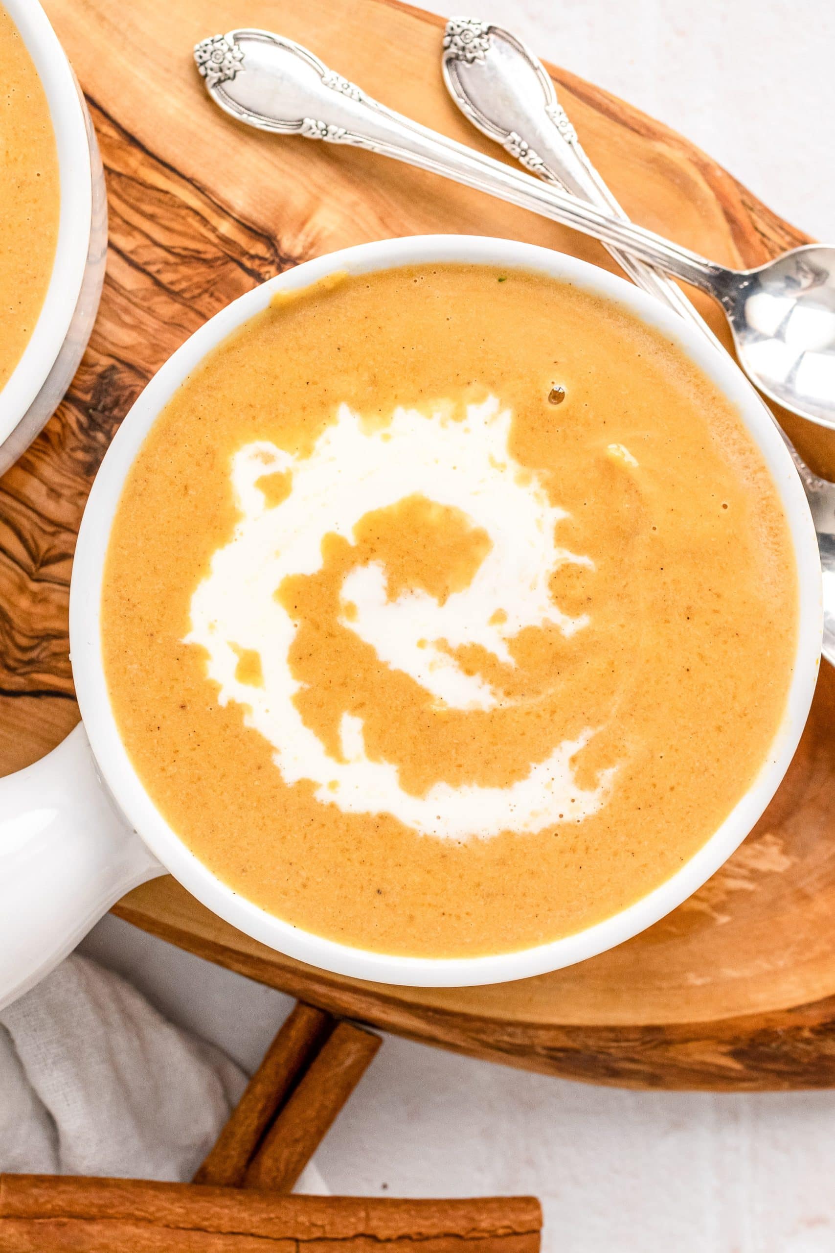 Overhead look at bowl of butternut squash soup with swirl of cream on top. Two serving spoons and two cinnamon sticks off to the side for garnish.