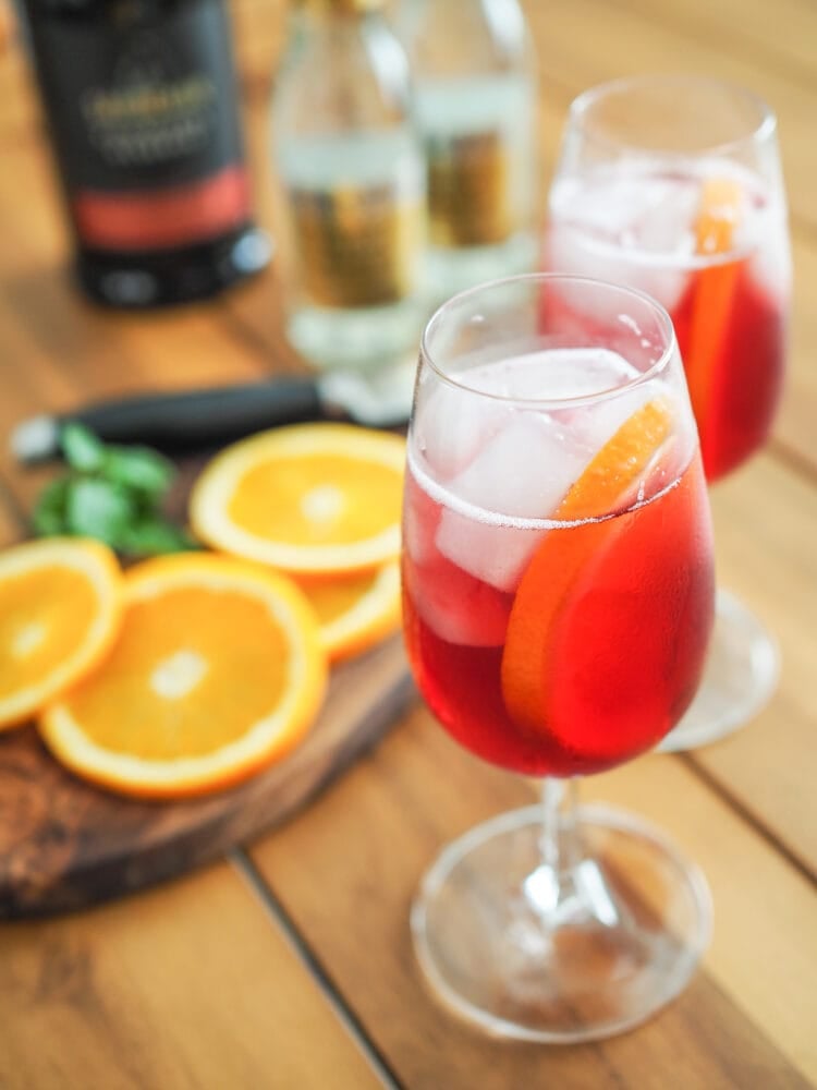 Two port and tonic drinks with orange garnish on a cutting board and a bottle of Port wine and bottle of tonic water in the background.