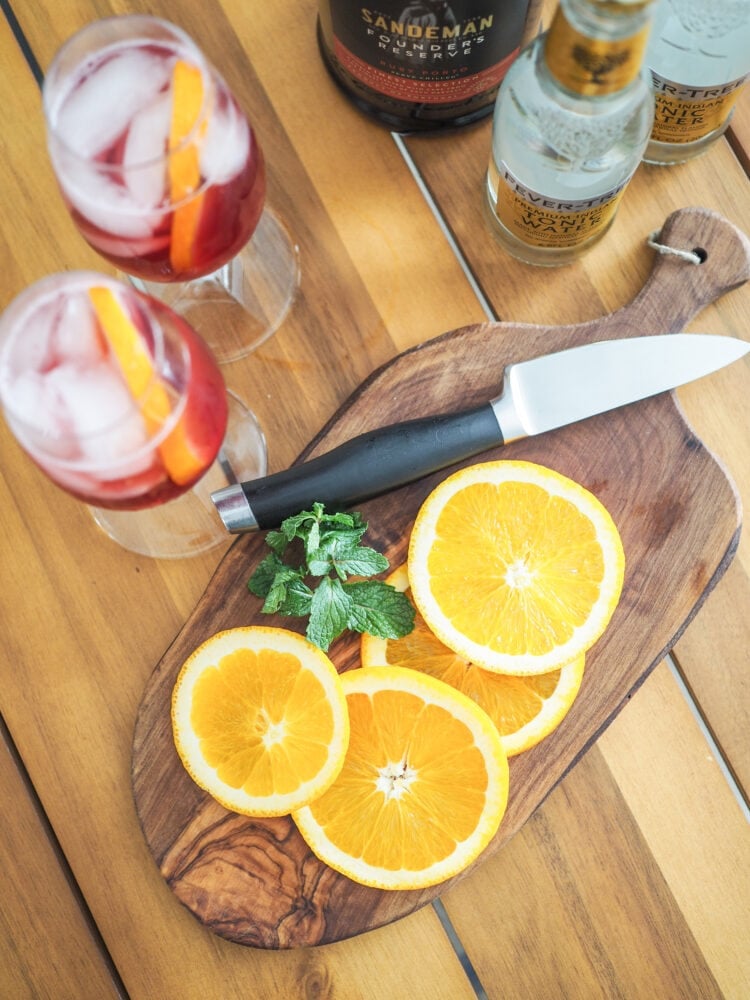 Overhead shot of Two port and tonic drinks with orange garnish on a cutting board and a bottle of Port wine and bottle of tonic water in the background.