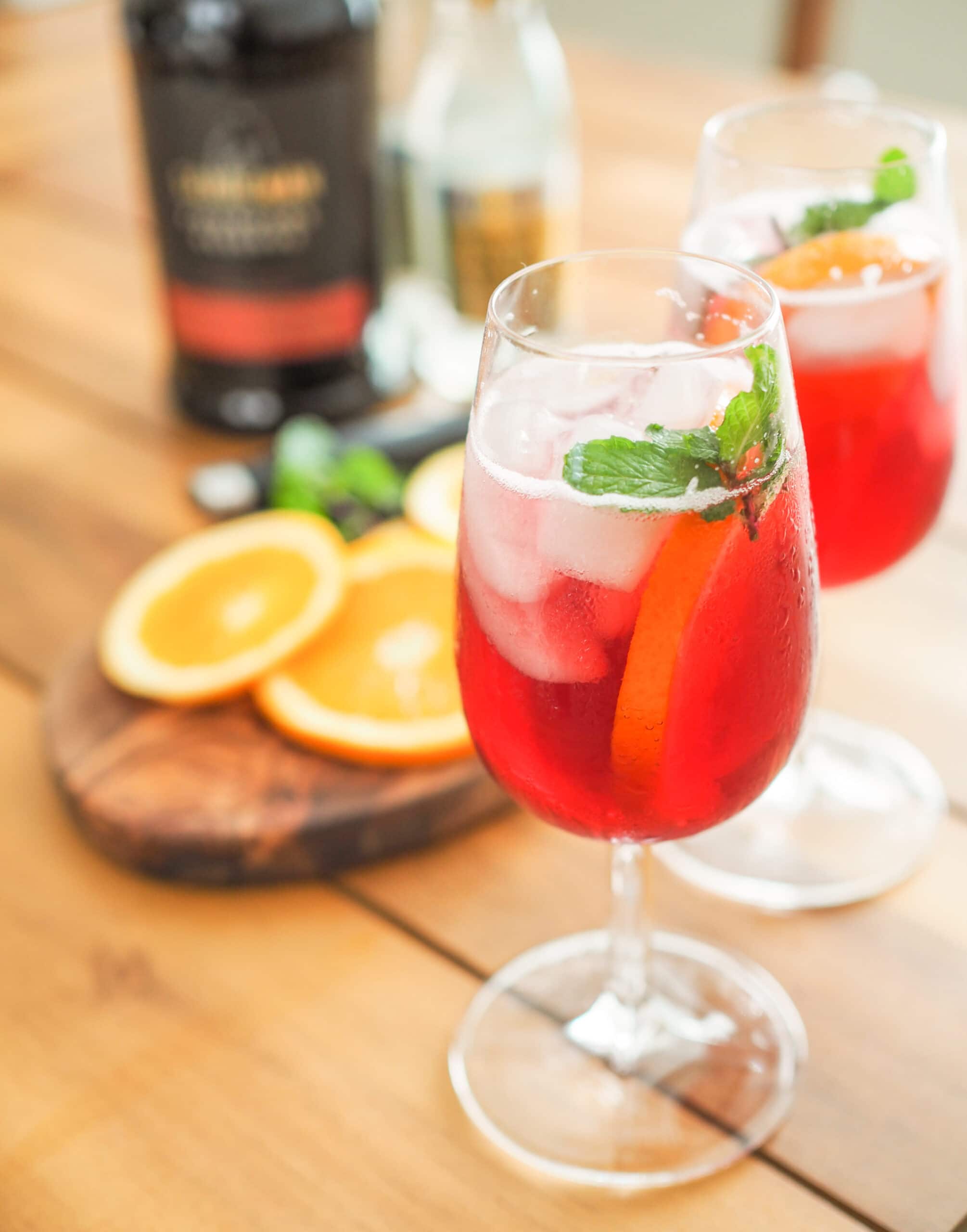 Two port and tonic drinks with orange garnish on a cutting board and a bottle of Port wine and bottle of tonic water in the background.