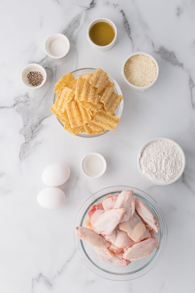 Overhead shot of ingredients to make crispy baked chicken wings including bowl of sunchips, pepper, salt, green tobasco, parmesan cheese, flour, eggs, wings, and water.