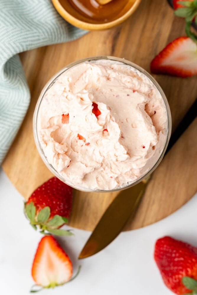 Overhead shot of small glass mason jar filled with whipped strawberry butter. It is resting on a cutting board with fresh cut strawberries and a small bowl with honey and a drizzle stick to the side.