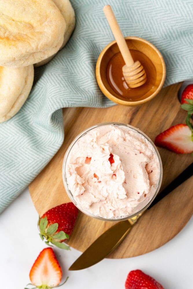 Overhead shot of small glass mason jar filled with whipped strawberry butter. It is resting on a cutting board with fresh cut strawberries and a small bowl with honey and a drizzle stick to the side.