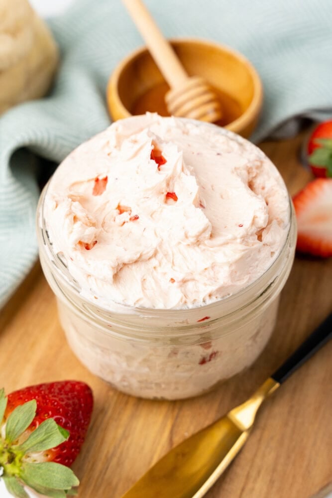 small glass mason jar filled with whipped strawberry butter. It is resting on a cutting board with fresh cut strawberries and a small bowl with honey and a drizzle stick to the side.