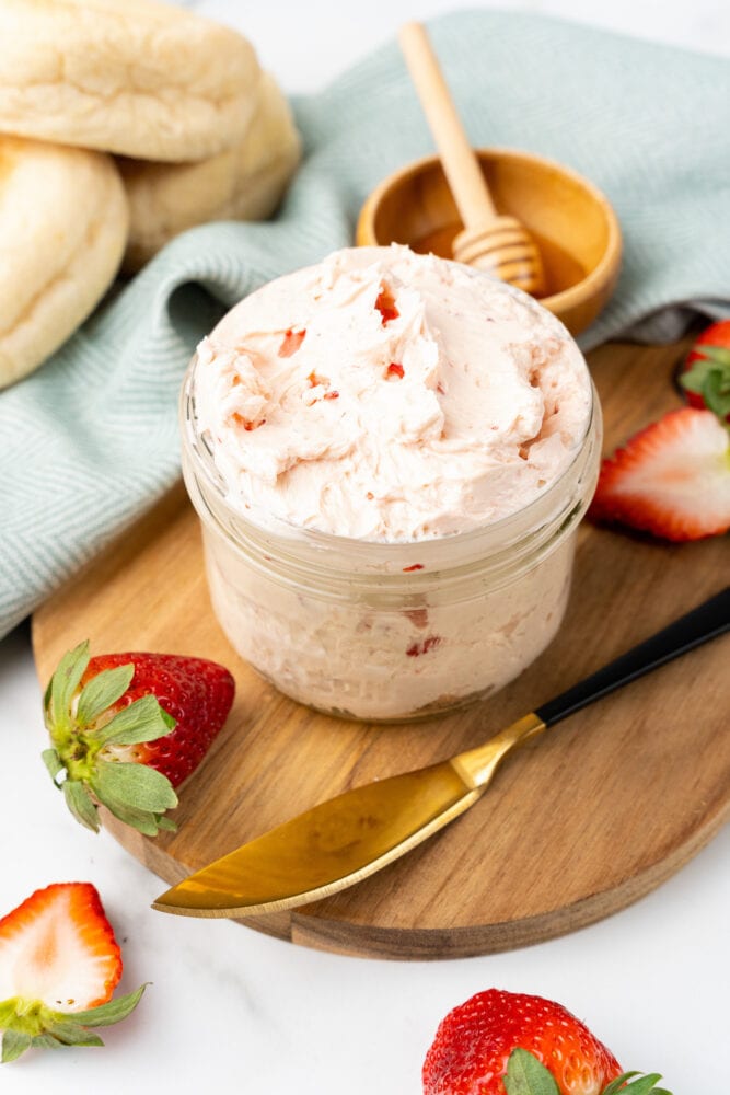 small glass mason jar filled with whipped strawberry butter. It is resting on a cutting board with fresh cut strawberries and a small bowl with honey and a drizzle stick to the side.