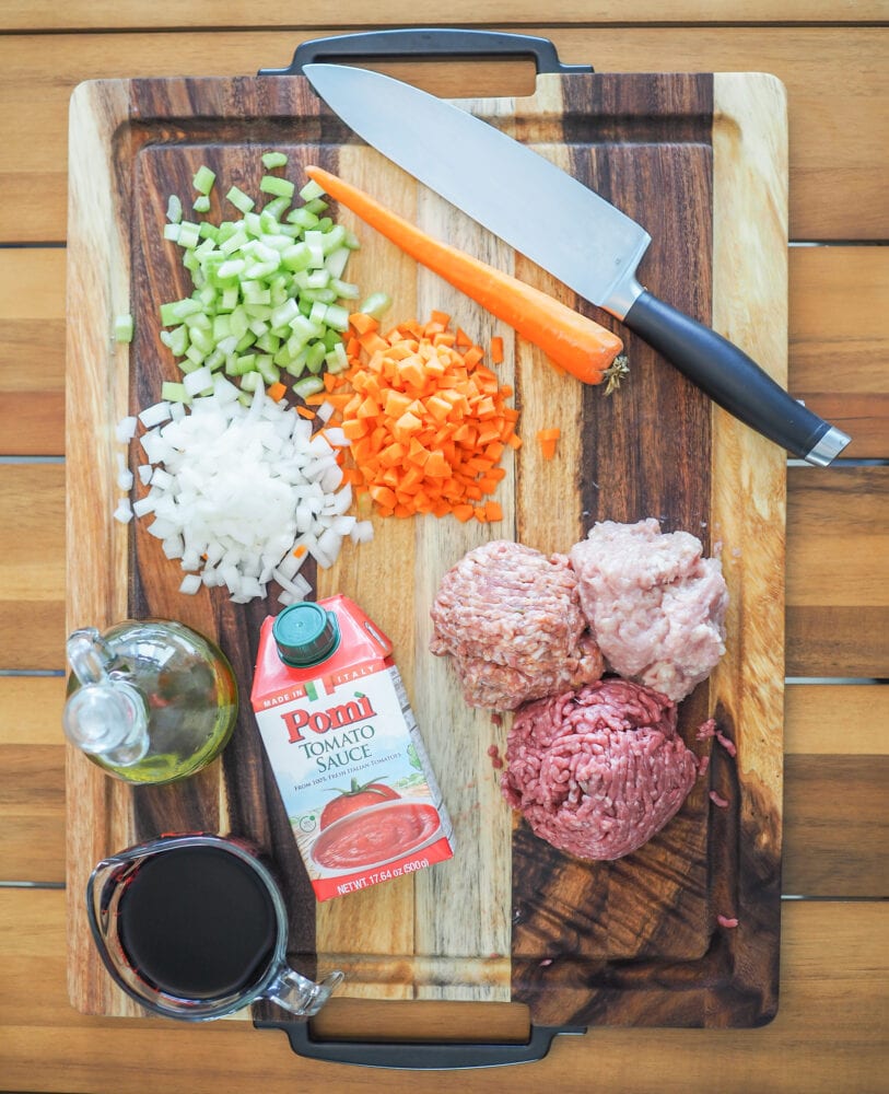 Overhead shot of cutting board with Bolognese sauce ingredients including: red wine, olive oil, tomato sauce, diced onions, diced celery, diced carrots, and three different ground meats include pork, beef, and sausage. 