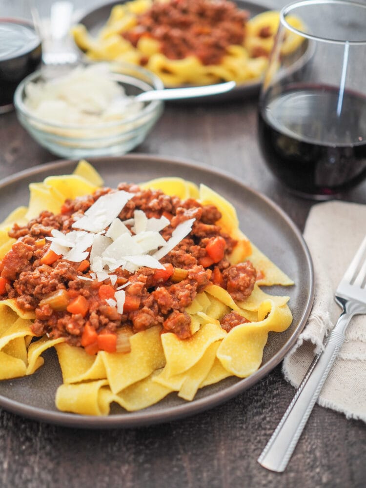 Pasta Bolognese made with an egg pappardelle noodle and topped with shaved Parmesan and served with a glass fo red wine.