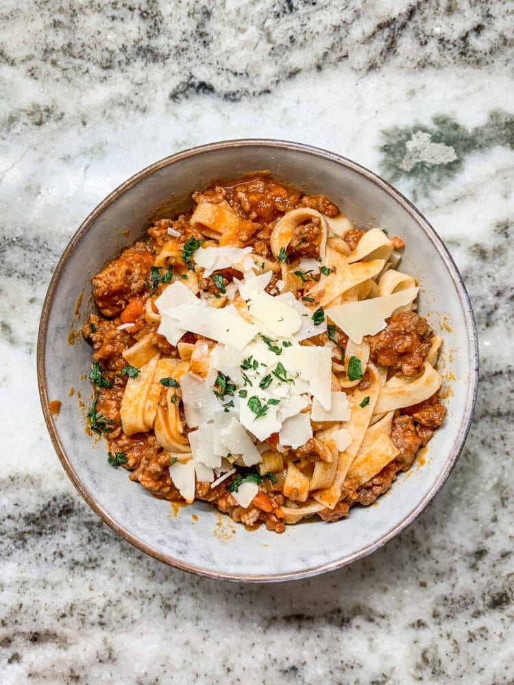 Overhead shot of a grey bowl of pasta bolognese made with fettuccini and topped with shaved Parmesan and parsley flakes.