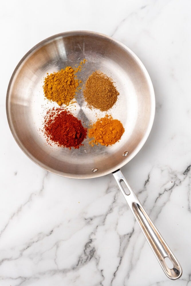 four spices in a dry stainless steel pan: turmeric, masala, chili, and ginger