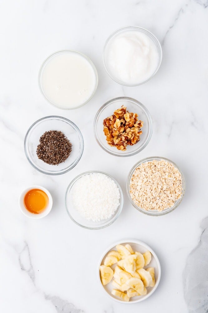 Ingredients for banana overnight oats including greek yogurt, coconut milk, chia seed, walnuts, oatmeal, coconut, honey, and sliced bananas.