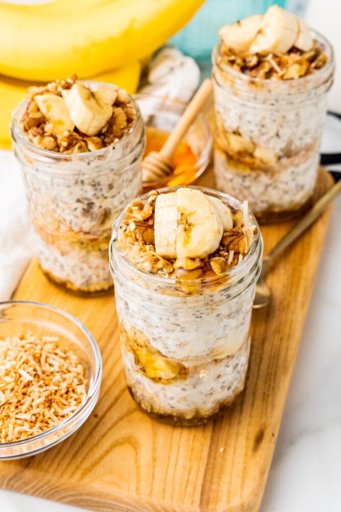 Three small mason jars layered with honey and banana overnight oats, topped with sliced bananas and walnuts, and a small dish of shredded toasted coconut to the side.