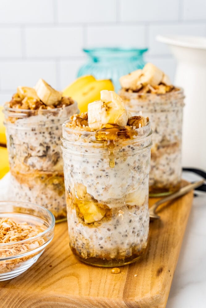 Three small mason jars layered with banana overnight oats, sliced bananas. Honey is being drizzled along the top.