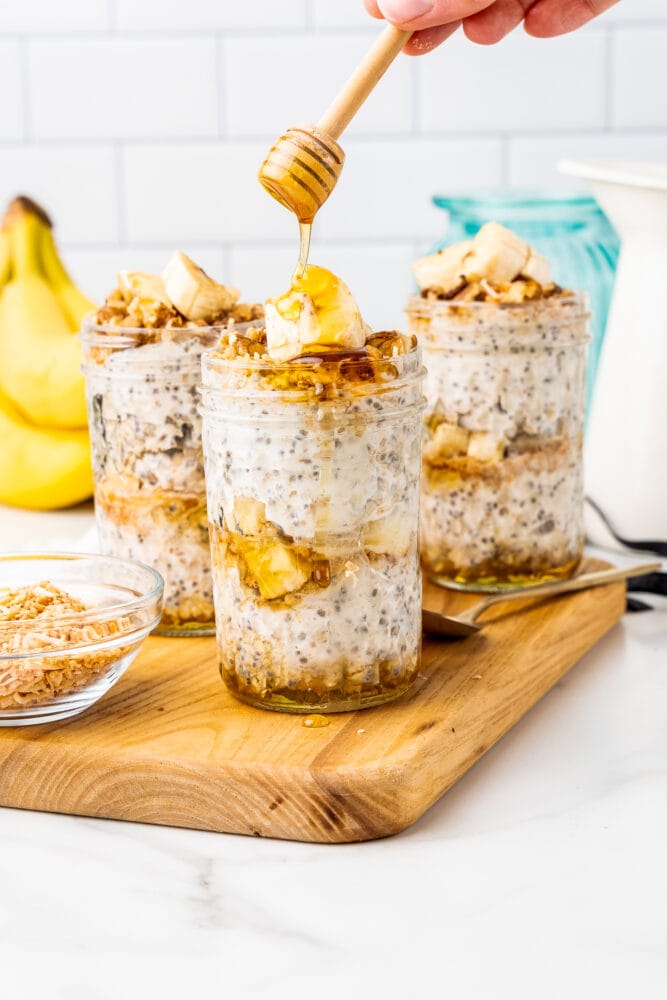Three small mason jars layered with banana overnight oats, sliced bananas. Honey is being drizzled along the top.