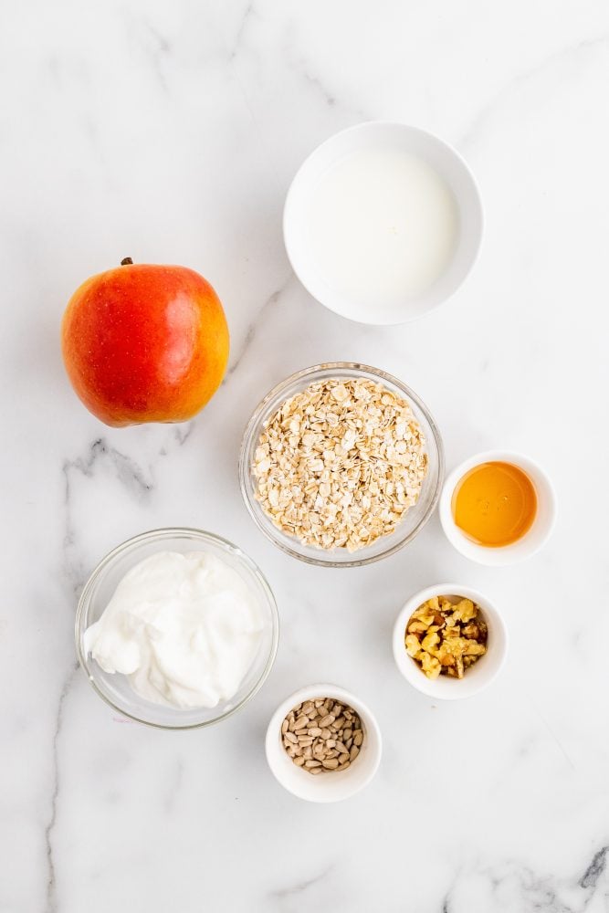 Overhead look at the ingredients needed for your homemade muesli recipe including a whole apple, rolled oats, milk, honey, yogurt, walnuts, and sunflower seeds.