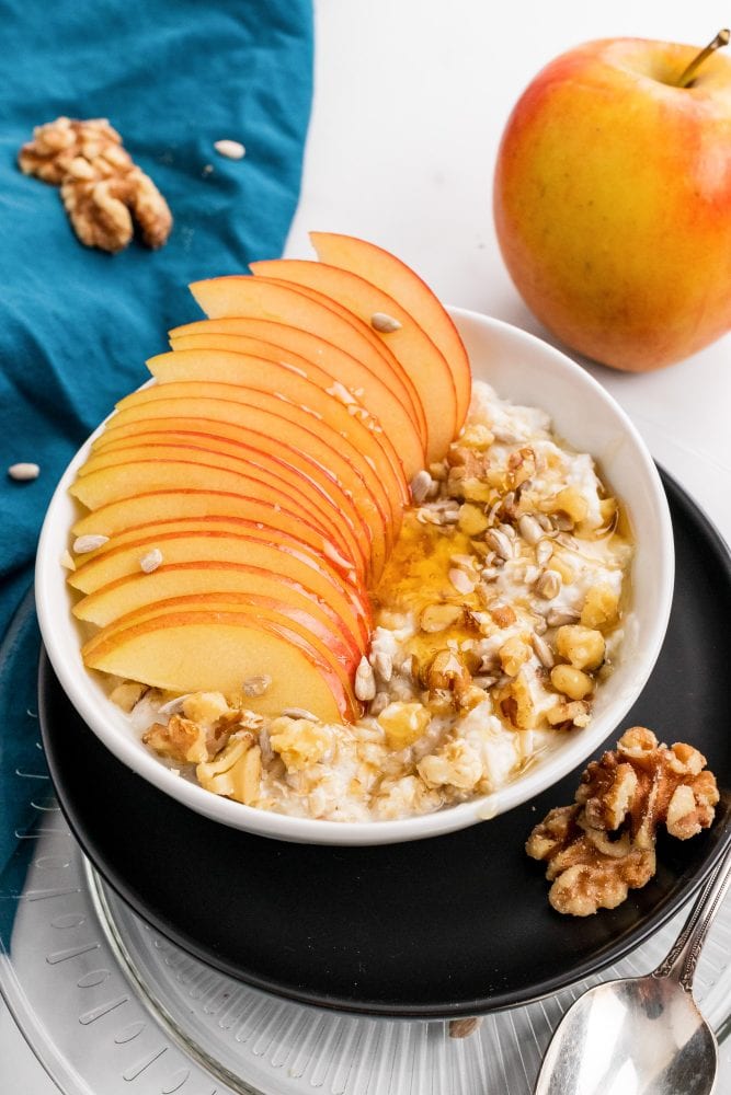 Overhead look at a bowl of muesli topped with walnuts and sliced apple, served in a white bowl over a black plate with a blue napkin and vintage style spoon.