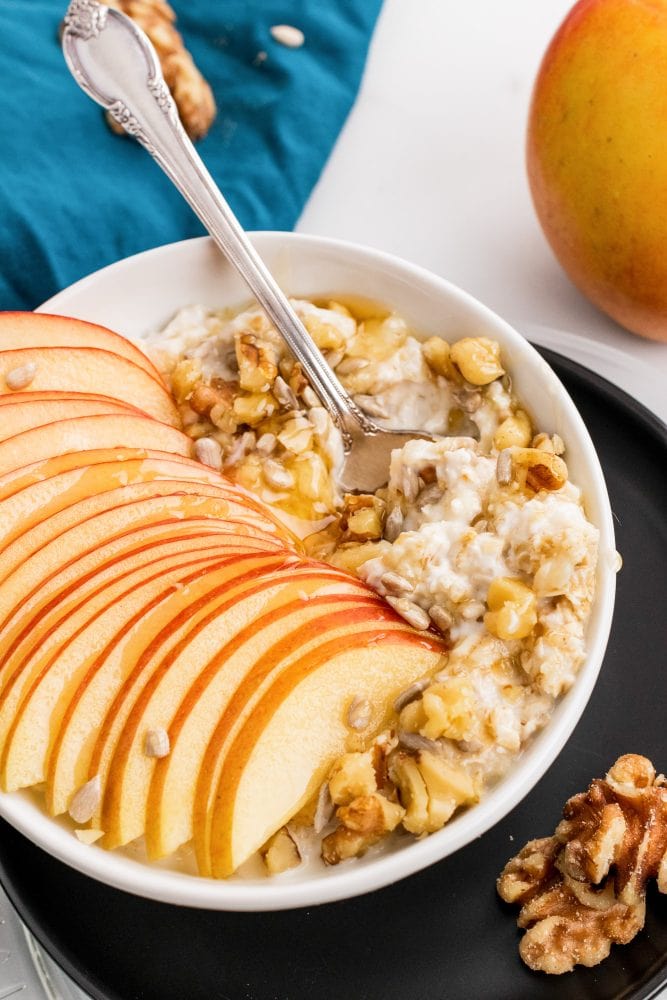 Overhead look at a bowl of muesli topped with walnuts and sliced apple, served in a white bowl over a black plate with a blue napkin and vintage style spoon.