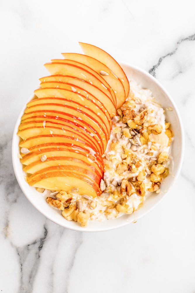 Overhead look at a white bowl of muesli topped with walnuts and sliced apple. Served on a white marble table.