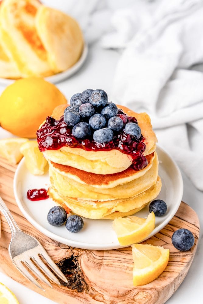 Stack of lemon ricotta blueberry pancakes on a white plate, covered in blueberry preserves and fresh blueberries. The plate is resting on a wood board with a fork to the left of the plate.