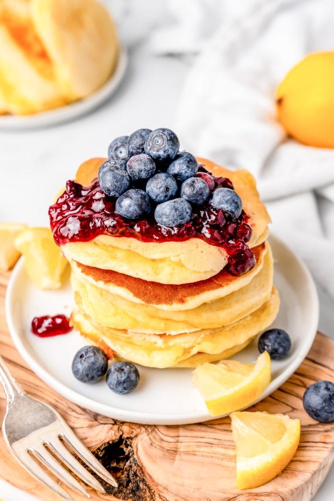 Stack of lemon ricotta blueberry pancakes on a white plate, covered in blueberry preserves and fresh blueberries. The plate is resting on a wood board with a fork to the left of the plate.