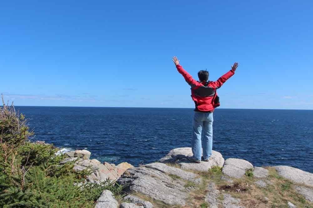 Photo of the back of Pete, his hands raised in the air, standing on a boulder overlooking the ocean at a lookout point.