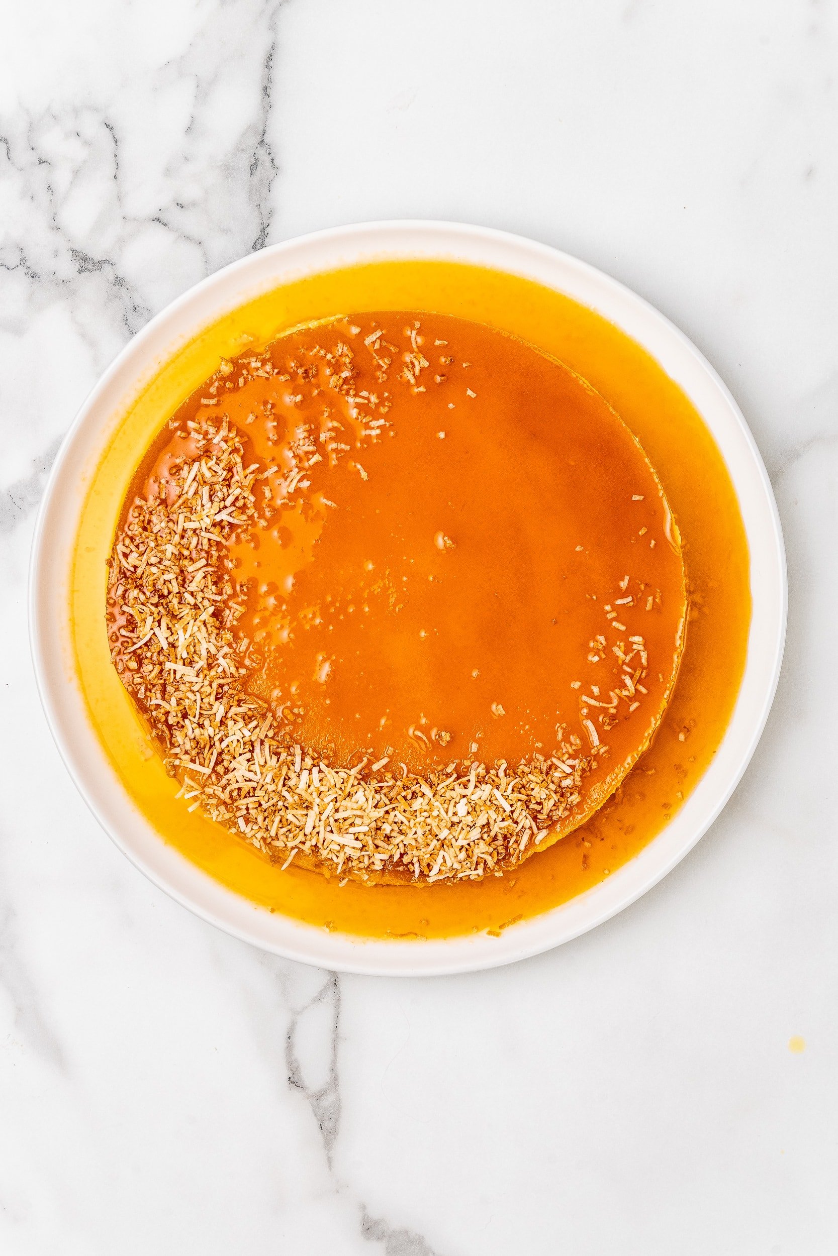 Overhead look at whole coconut flan topped with toasted stredded coconut and caramel on a white serving plate.