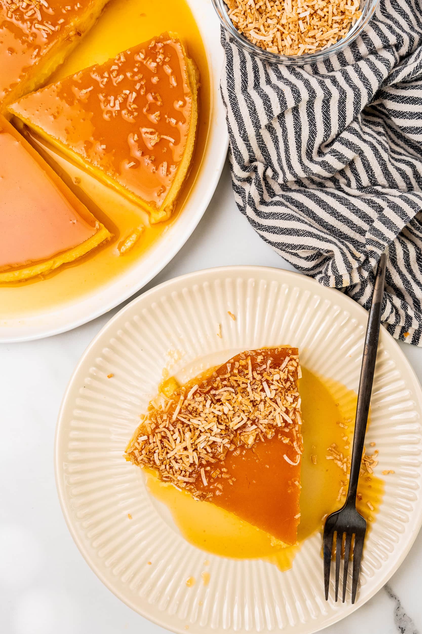 Large serving plate with coconut flan cut into pie shaped slices, with one slice on a small plate with a black fork. There's a black and white striped towel and toasted shredded coconut in a small bowl to the side.