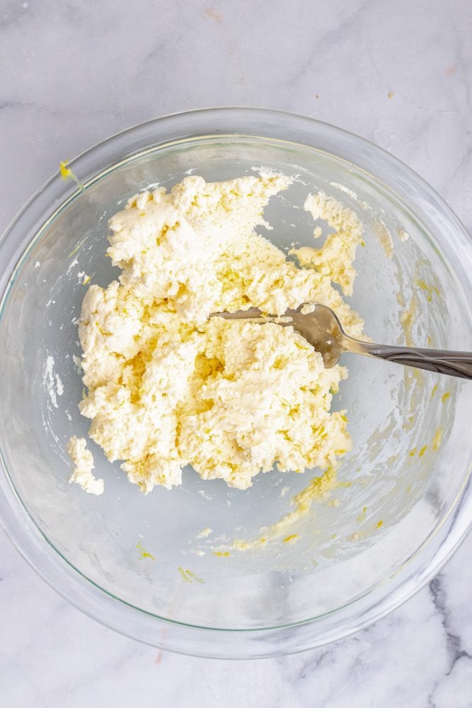 Overhead look at a clear glass bowl with mascarpone cheese mixed with lemon zest and juice.