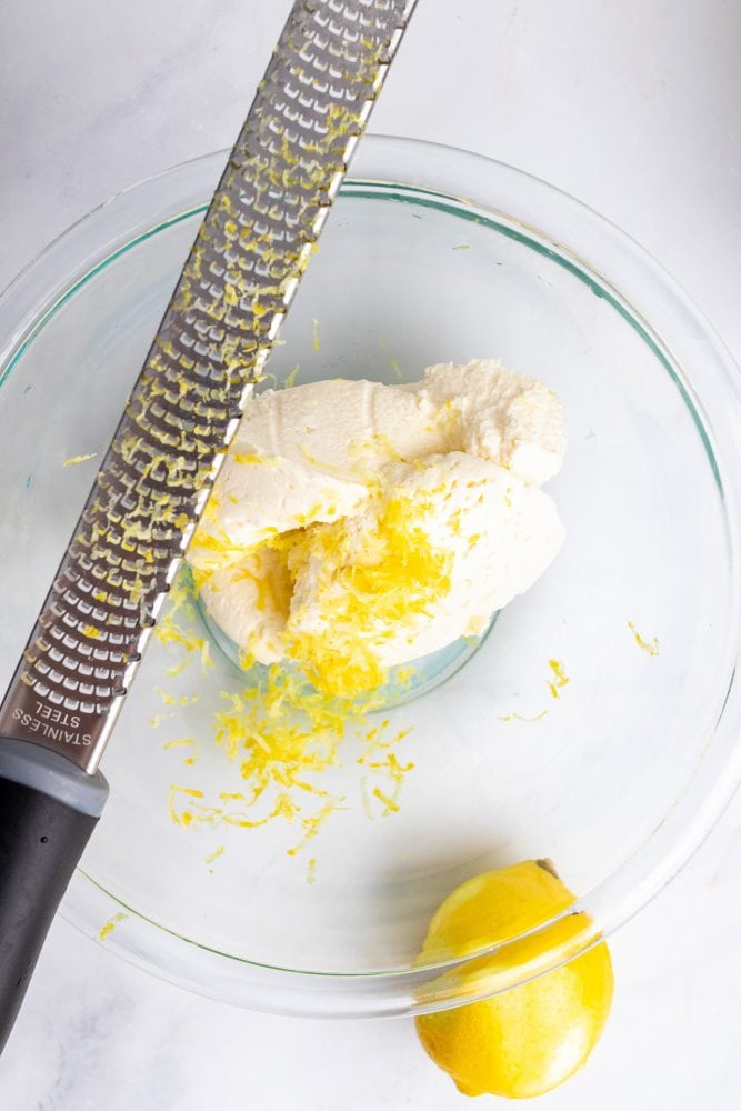 Overhead look at mascarpone cheese in a clear glass bowl with lemon zest.