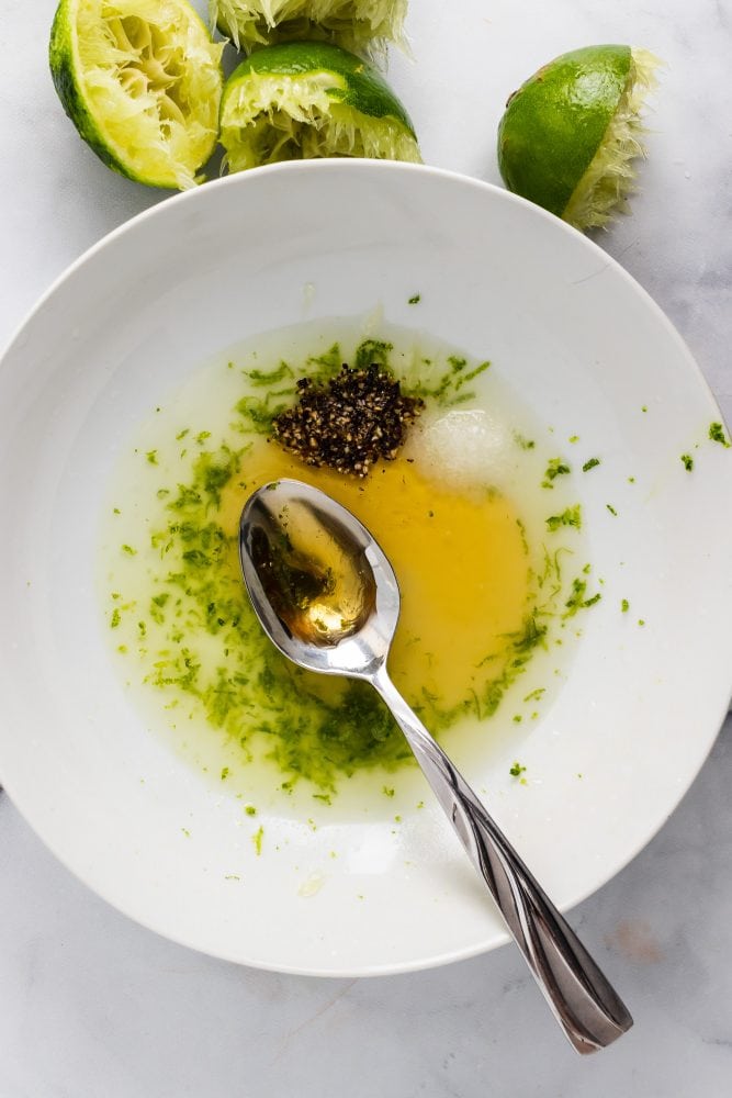 Overhead look at bowl filled with dressing ingredients (honey, lime zest, salt and pepper) ready to be whisked.