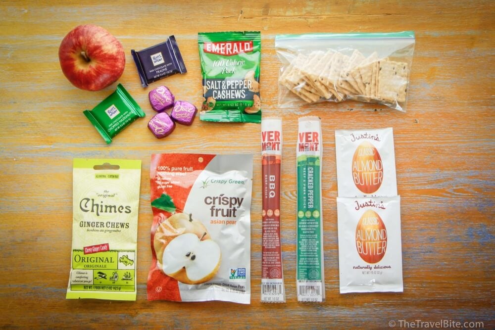 Road Trip Essentials: Overhead shot of snacks including an apple, Chimes ginger chews, chocolate, dehydrated pears, cashews, beef jerky, crackers, and almond butter.